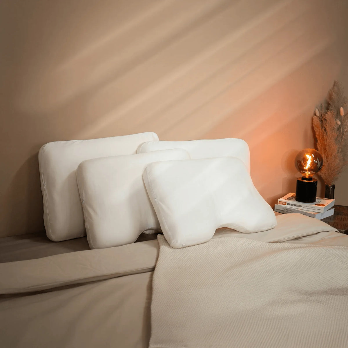 Drei weiße Vorteilsset: Baumwoll-Kissenbezug-Kissen von Liebscher & Bracht liegen auf einem ordentlich gemachten Bett mit beiger Bettwäsche, einer beigen Überwurfdecke und einem Nachttisch mit Büchern, einer Lampe und getrocknetem Pampasgras.