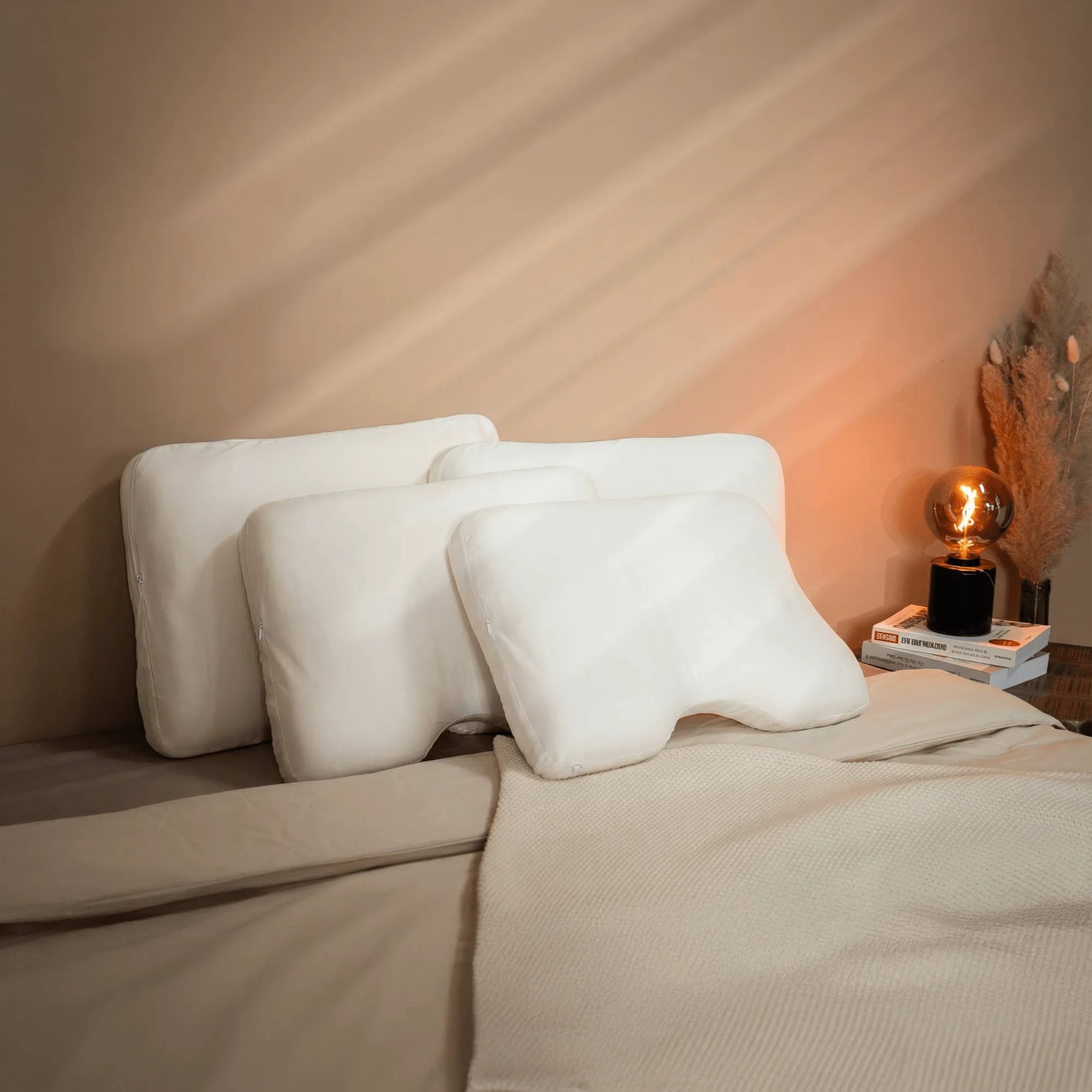 Drei weiße Vorteilsset: Baumwoll-Kissenbezug-Kissen von Liebscher & Bracht liegen auf einem ordentlich gemachten Bett mit beiger Bettwäsche, einer beigen Überwurfdecke und einem Nachttisch mit Büchern, einer Lampe und getrocknetem Pampasgras.