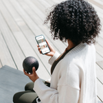 Eine Person mit lockigem Haar, weißem Pullover und grünen Leggings sitzt im Freien auf einer Yogamatte und kontrolliert ihre Übungen mit der Liebscher & Bracht App, während sie einen schwarzen Massageball von Liebscher & Bracht in der Hand hält.