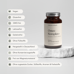 Eine braune Glasflasche von Liebscher & Bracht Osteo Komplex+ für 2 Monate mit Vitamin D steht auf einem weißen Stein. Icons auf der linken Seite heben Merkmale hervor: vegan, umweltfreundlich, glutenfrei, ohne Gentechnik, laktosefrei und mehr.