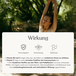 Eine Frau steht im Freien, die Augen geschlossen, die Arme über den Kopf erhoben. Im Vordergrund hebt Zell Komplex+ von Liebscher & Bracht hervor, wie seine Nährstoffe, wie Vitamin C, die Immunität und den Zellschutz für 2 Monate unterstützen.