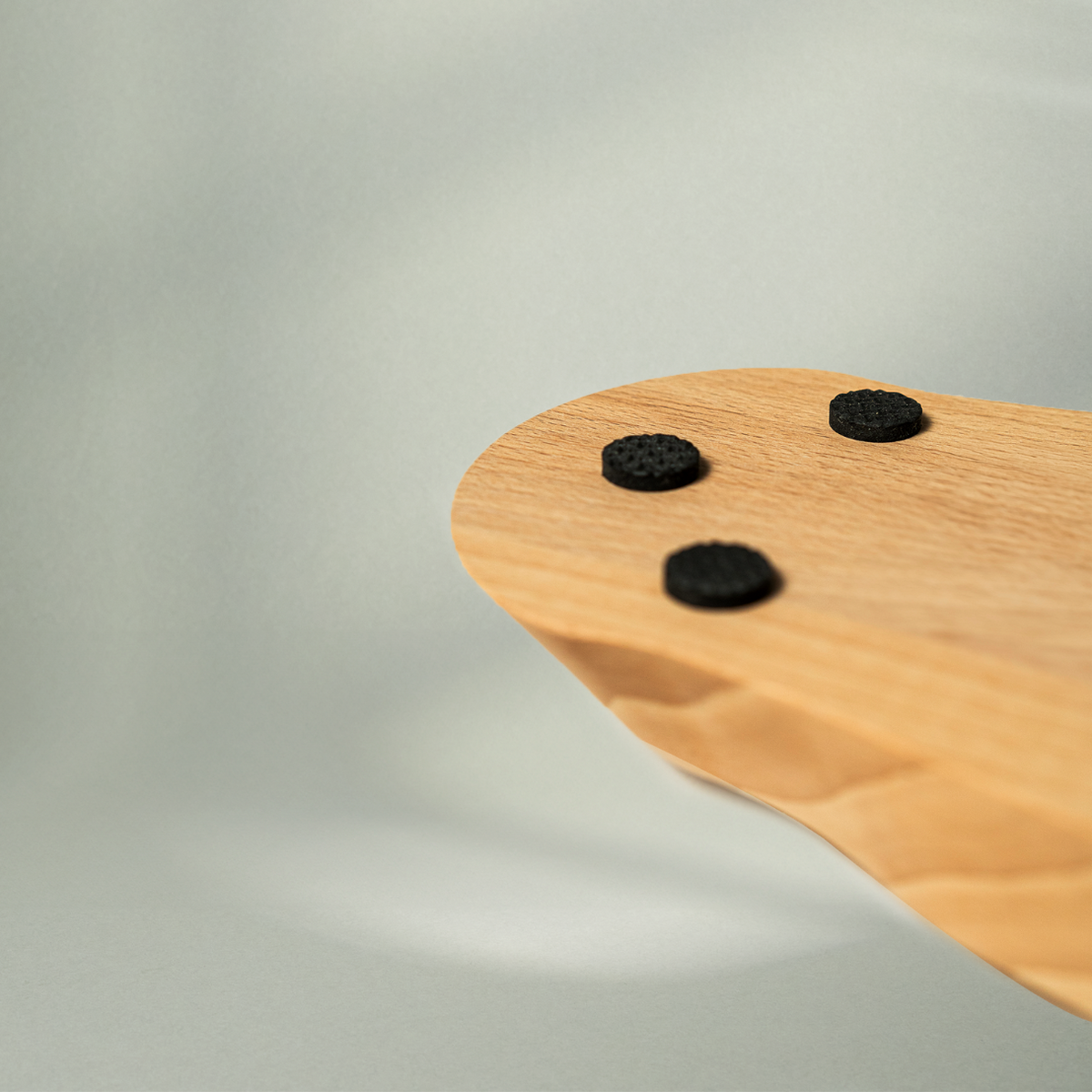 Eine Nahaufnahme des Liebscher & Bracht Holz-Sockels für den Nackenretter, ein geschwungenes Holz-Accessoire mit drei runden schwarzen Pads, vor einem weichen hellgrauen Hintergrund.