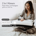 Eine Frau im Pyjama sitzt auf einem Bett und begutachtet das Liebscher & Bracht Aktiv-Set (1 Jahr App + Kissen). Der deutsche Text hebt die Eigenschaften des Kissens hervor: eine weiche Seite, eine mittelharte Seite und einen verstellbaren Bezug.