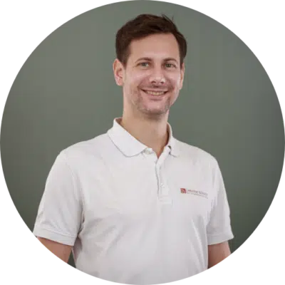 Ein Mann mit kurzen braunen Haaren lächelt in die Kamera. Er trägt ein weißes Poloshirt mit einem kleinen Logo auf der Brust und steht vor einem schlichten grauen Hintergrund.