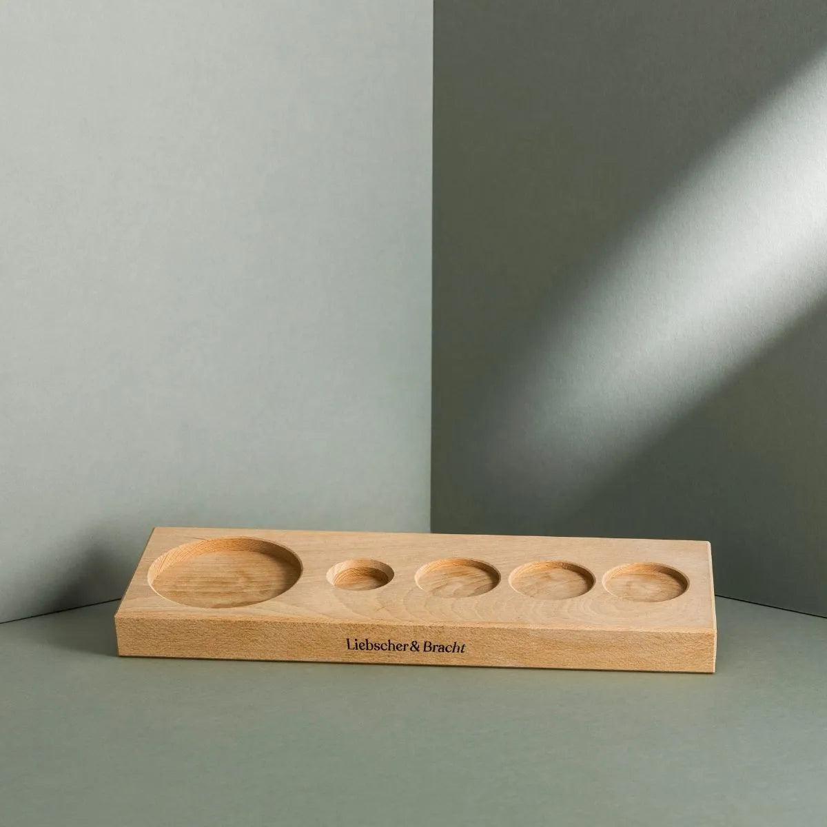 Das Aufbewahrungsbrett von Liebscher & Bracht ist ein rechteckiges Holztablett mit fünf runden, abgestuften Vertiefungen zur Aufnahme eines Drücker-Sets, das auf einer grauen Fläche mit passendem Hintergrund steht und den Markennamen trägt.