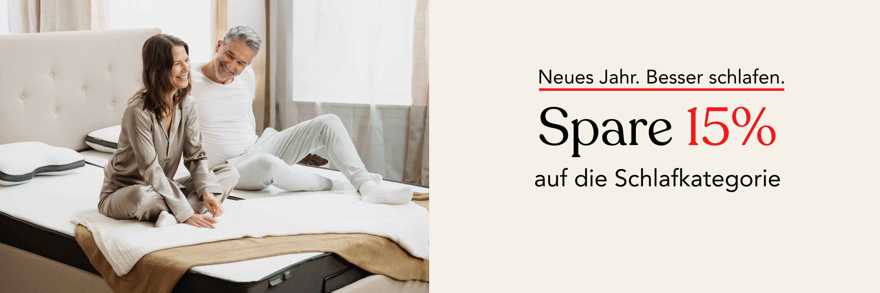 Ein lächelndes Paar im Schlafanzug sitzt auf einem sauber gemachten Bett in einem hellen Schlafzimmer. Der Text neben ihnen lautet: Neues Jahr. Besser schlafen. Spare 15% auf die Schlafkategorie.