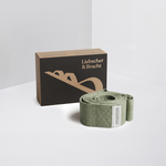 Neben einem aufgerollten grünen Gurt mit weißem Etikett steht eine „Der Schulterretter“-Box von Liebscher & Bracht – ideal für Übungen zur Linderung von Schulterschmerzen –, die auf einer weißen Fläche vor einem minimalistischen Hintergrund präsentiert wird.