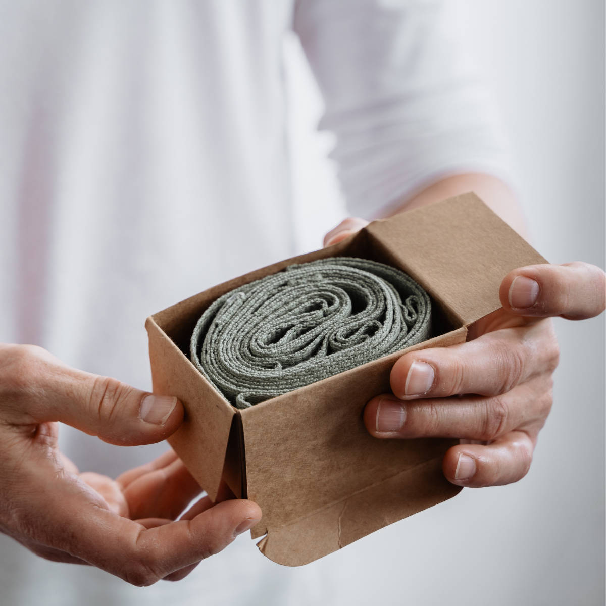 Eine Person hält vor einem einfarbigen Hintergrund einen offenen braunen Karton in der Hand, in dem sich ein aufgerolltes graues Textilprodukt befindet – „Der Schulterretter“ von Liebscher & Bracht.
