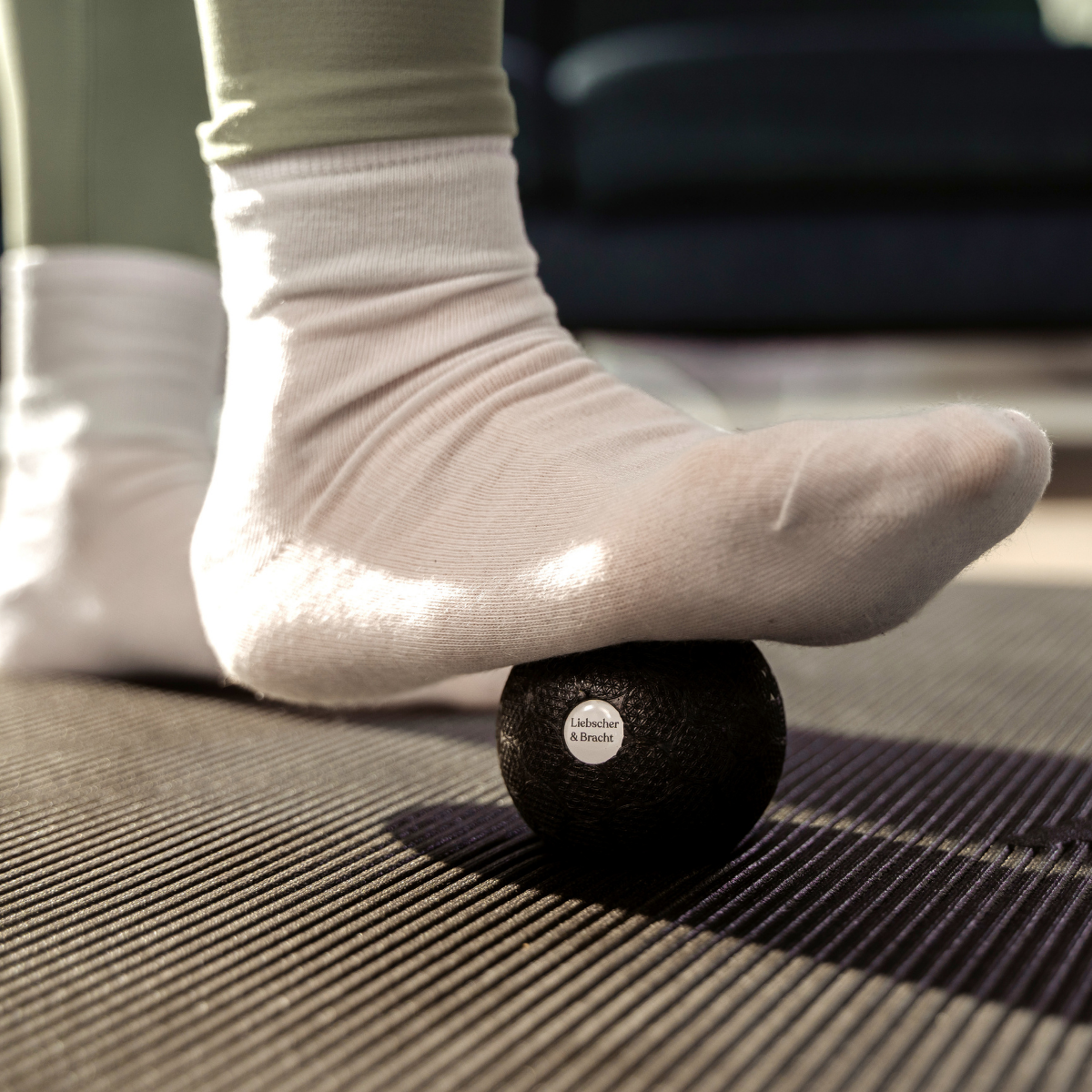 Jemand in weißen Socken nutzt das Faszien-Rollmassage-Set von Liebscher & Bracht und rollt seinen Fuß auf einem schwarzen Massageball über eine strukturierte Matte – zu Hause, zur Entspannung oder um durch Fußmassage oder Bewegungstherapie die Beweglichkeit zu verbessern.