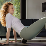 Eine Frau mit lockigem rotem Haar wendet das Faszien-Rollmassage-Set von Liebscher & Bracht auf einer Yogamatte in einem Innenraum unter ihrem unteren Rücken an, um Entspannung und Beweglichkeit zu fördern; im Hintergrund sind ein Sofa und ein Fenster zu sehen.