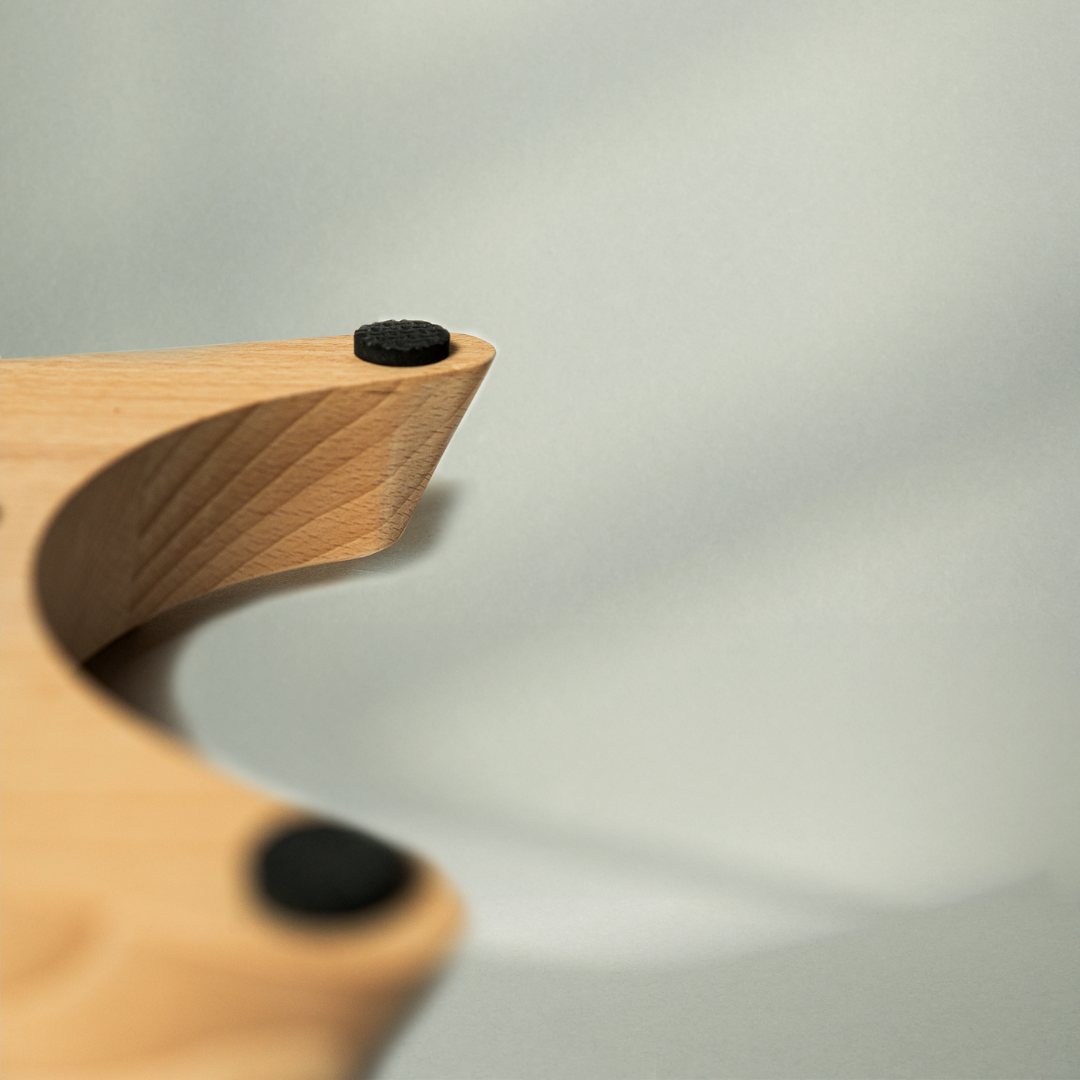 Eine Nahaufnahme des Liebscher & Bracht Holz-Sockels für den Nackenretter aus hellem Holz mit sanften Rundungen und schwarzen, kreisförmigen Pads auf der Oberseite vor einem weichen, neutralen Hintergrund mit sanften Schatten.