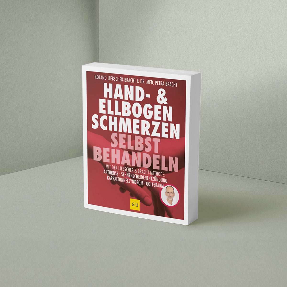 Das Buch "Hand & Ellenbogen Schmerzen selbst behandeln" von Liebscher & Bracht steht aufrecht auf einer hellgrauen Fläche vor einer hellgrauen Wand. Der rote Einband ist mit weißer und gelber Schrift sowie einem kleinen runden Foto einer Person versehen.