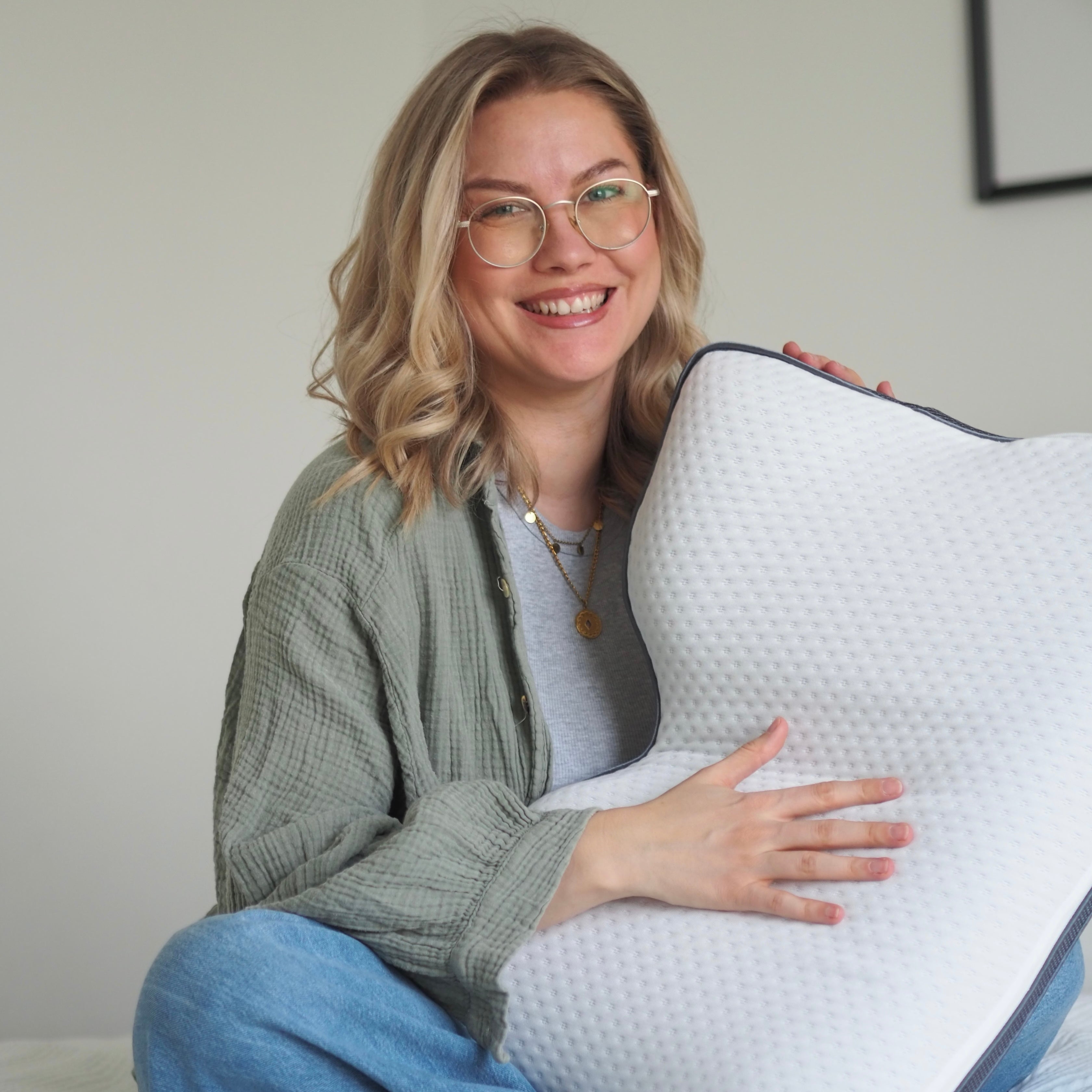 Eine lächelnde Frau mit blondem Haar und Brille hält ein großes, weißes Memory Foam-Kissen mit strukturiertem Bezug. Sie trägt ein grünes Hemd über einem grauen Oberteil und sitzt vor einem schlichten Hintergrund.
