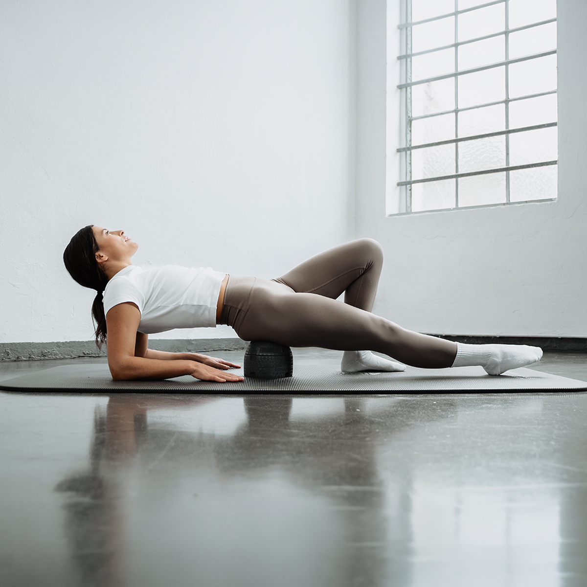 Eine Frau in Sportkleidung liegt auf dem Rücken auf einer Yogamatte und nutzt den Liebscher & Bracht ISG-Ischias-Retter unter ihren Hüften zur Linderung von ISG-Beschwerden in einem hellen Raum mit großen Glasbausteinfenstern.