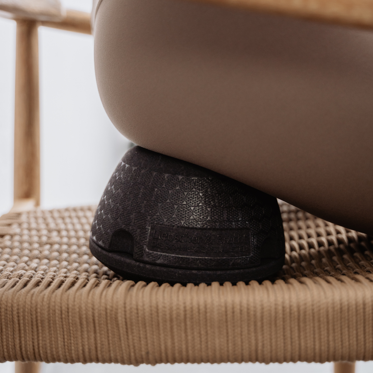 Eine Person sitzt auf einem geflochtenen Stuhl und hat das schwarze, ergonomische Kissen „Der ISG-Ischias-Retter“ von Liebscher & Bracht unter dem Oberschenkel – ideal für eine gezielte Schmerztherapie und zur Linderung von Hüftschmerzen oder ISG-Beschwerden.