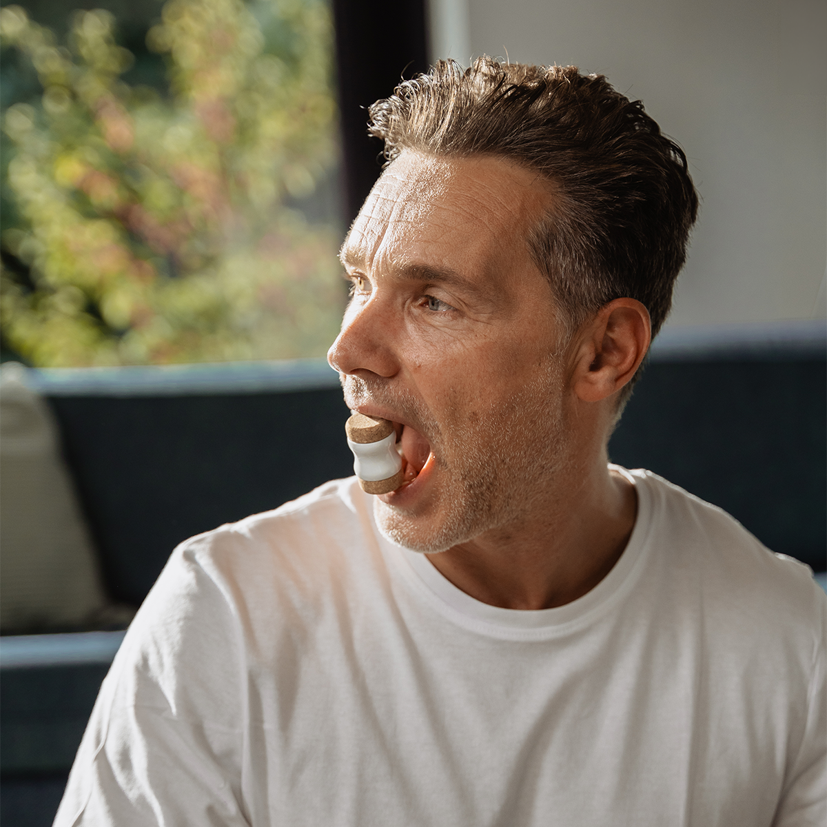 Ein Mann mit kurzen Haaren und Bartstoppeln, der ein weißes T-Shirt trägt, sitzt in einem Raum und benutzt den „Kieferretter“ von Liebscher & Bracht gegen Zähneknirschen; sein Mund ist geöffnet, während seitlich natürliches Licht hereinscheint.