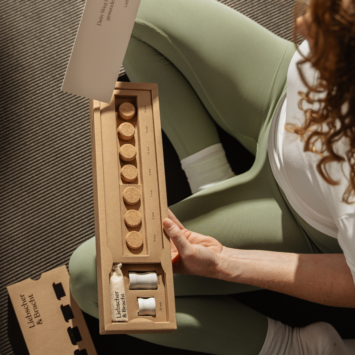 Eine Person in grünen Leggings und einem weißen Hemd sitzt auf dem Boden und hält eine geöffnete Schachtel „Der Kieferretter“ von Liebscher & Bracht in der Hand, die sechs runde Tabletten und zwei kleine Fläschchen enthält und vermutlich zur Behandlung von Zähneknirschen oder CMD-Symptomen gedacht ist.