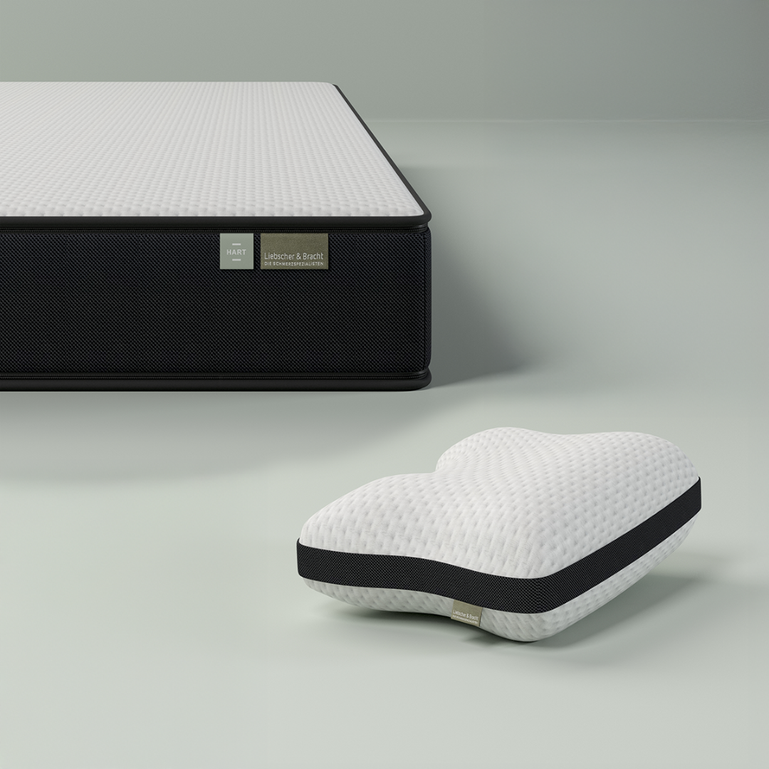 Das Liebscher & Bracht Schlafset: Matratze + Kissen besteht aus einer modernen Matratze mit weißer, strukturierter Oberseite und schwarzen Seiten, gepaart mit einem weißen, herzförmigen Kissen mit einem schwarzen Streifen im Vordergrund.