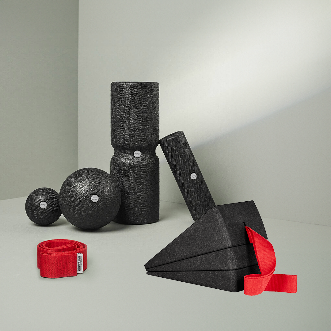 Das Dein Knie-Paket von Liebscher & Bracht besteht aus schwarzen, strukturierten Schaumstoffwerkzeugen - zwei Faszienrollen, zwei Bälle, zwei Dreiecksblöcke und eine rote Trittschlaufe - die auf einer hellgrauen Fläche vor einem hellen Hintergrund angeordnet sind.