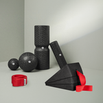 Das Dein Knie-Paket von Liebscher & Bracht besteht aus schwarzen, strukturierten Schaumstoffwerkzeugen - zwei Faszienrollen, zwei Bälle, zwei Dreiecksblöcke und eine rote Trittschlaufe - die auf einer hellgrauen Fläche vor einem hellen Hintergrund angeordnet sind.