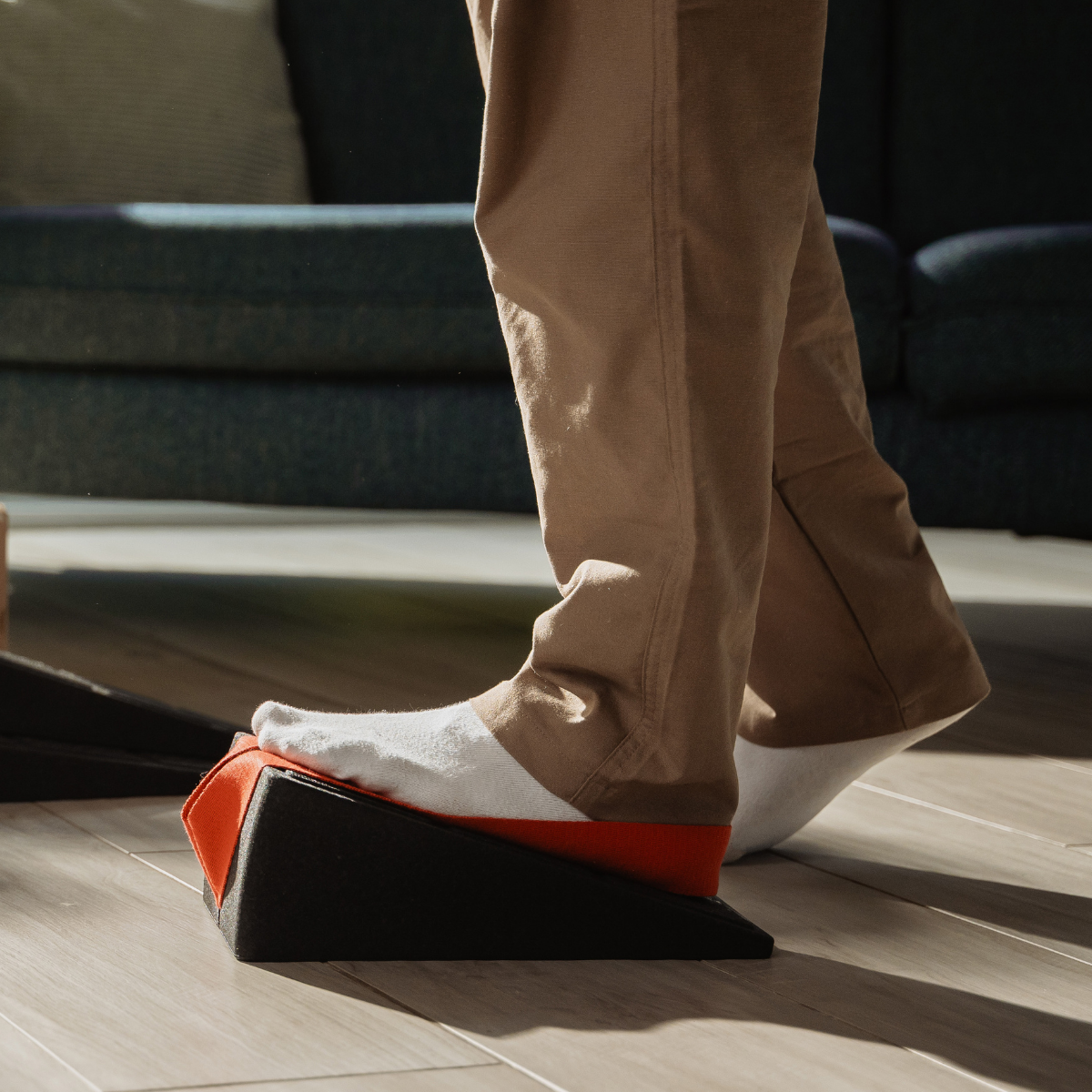 Eine Person in hellen Socken und beigen Hosen streckt ihren Fuß auf dem „Liebscher & Bracht Der Knieretter mit Trittschlaufe“ aus – einem schwarz-orangefarbenen Schaumstoffkeil –, der auf einem Holzboden steht, im Hintergrund ist ein dunkles Sofa zu sehen.