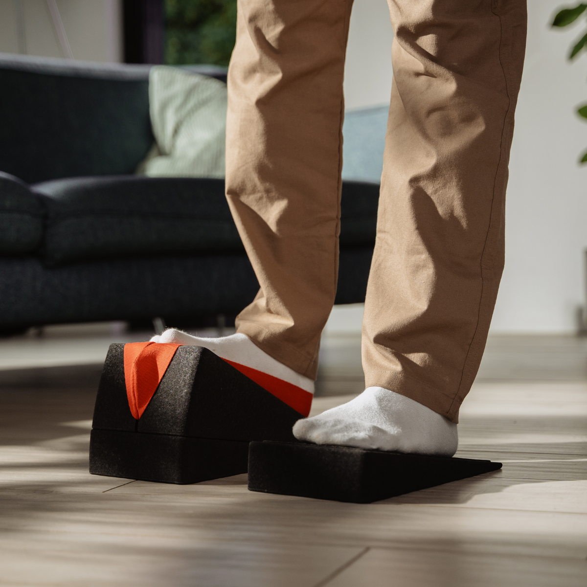 Eine Person steht auf zwei schwarzen Schaumstoffkeilen „Der Knieretter mit Trittschlaufe“ von Liebscher & Bracht und macht Übungen zur Linderung von Knieschmerzen in einem hellen Wohnzimmer, im Hintergrund ist ein Sofa zu sehen.