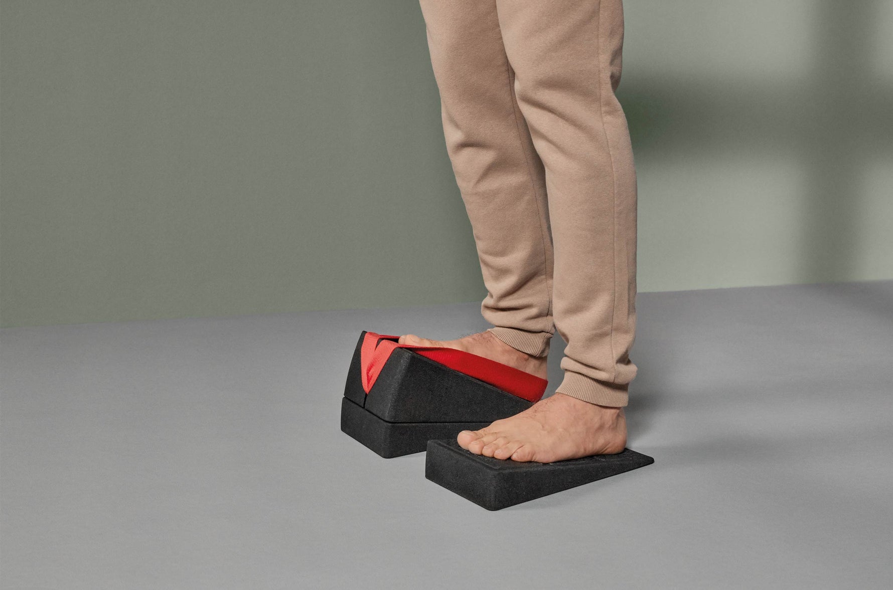 Eine Person, die eine beigefarbene Hose trägt, steht mit nackten Füßen auf zwei schwarzen Schaumstoff-Fußschalen; ein Fuß befindet sich in einer keilförmigen Vorrichtung mit roter Innenseite, während der andere auf einem schrägen Schaumstoffblock steht.