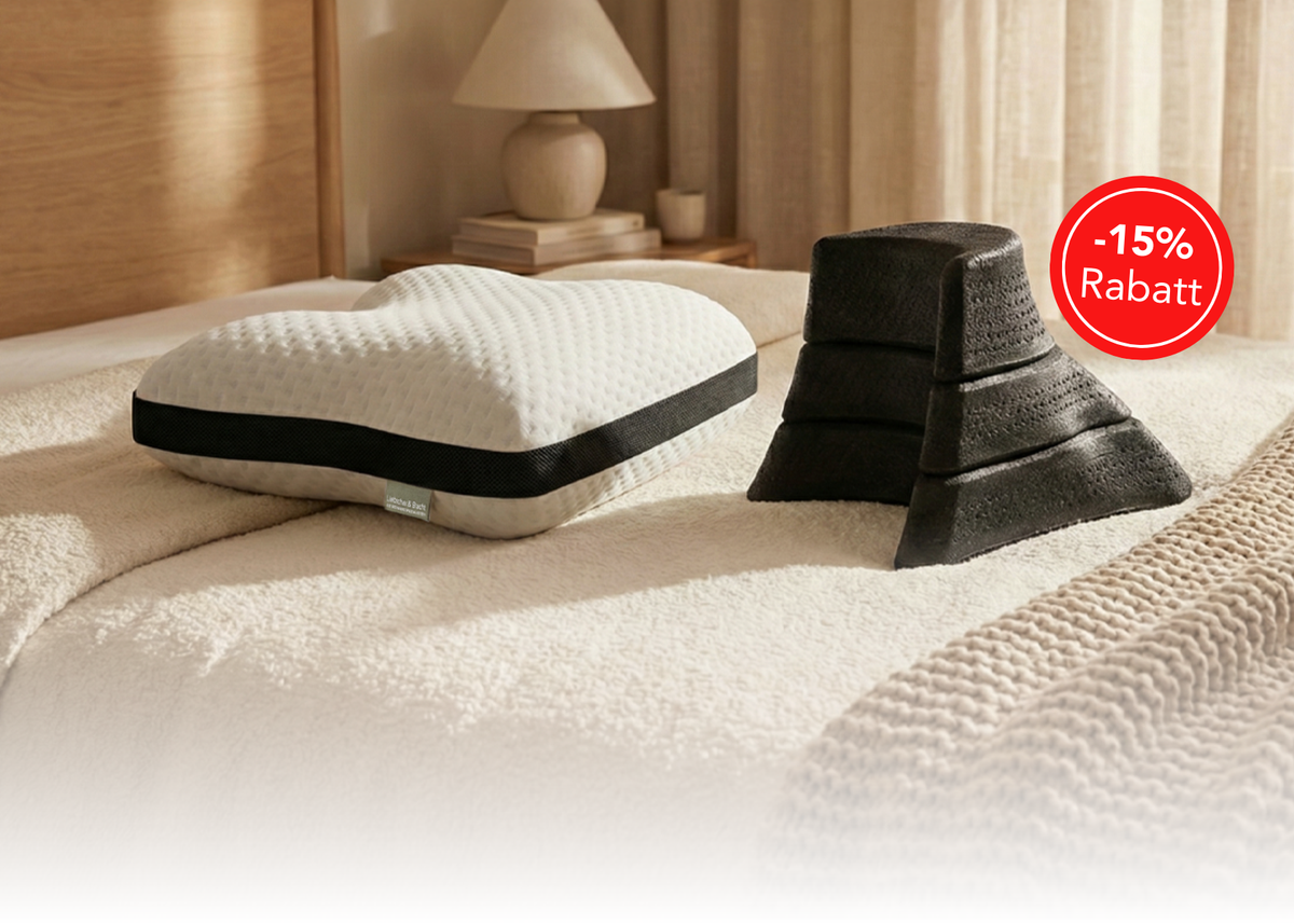 Ein herzförmiges weißes Kissen und ein Satz von drei gestapelten schwarzen Schaumstoffkeilen liegen auf einem ordentlich gemachten Bett. Ein roter Kreis in der Ecke zeigt -15% Rabatt an, was auf ein Rabattangebot hinweist.
