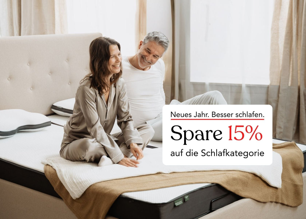 Ein lächelndes Paar sitzt auf einem modernen Bett in einem hellen Schlafzimmer. Ein Werbeschild in deutscher Sprache bietet einen Rabatt von 15 % auf Schlafprodukte für das neue Jahr.