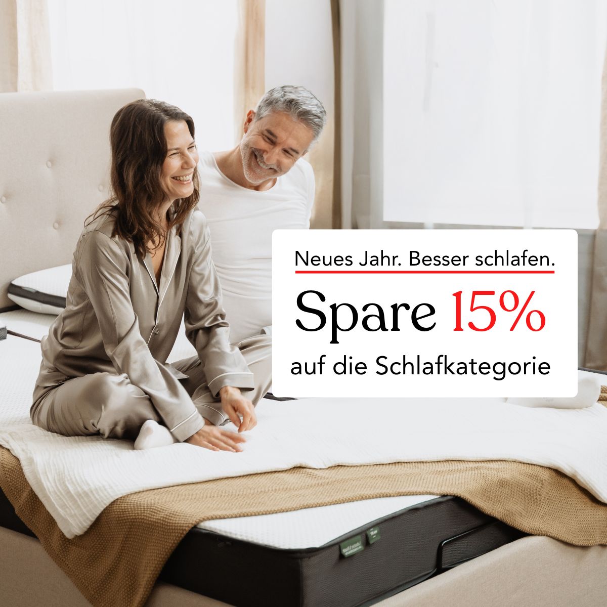 Ein lächelndes Paar sitzt auf einem sauber gemachten Bett in einem hellen Schlafzimmer. Der deutsche Text auf dem Bild wirbt für einen Rabatt von 15 % auf Schlafprodukte für das neue Jahr: Spare 15% auf die Schlafkategorie.
