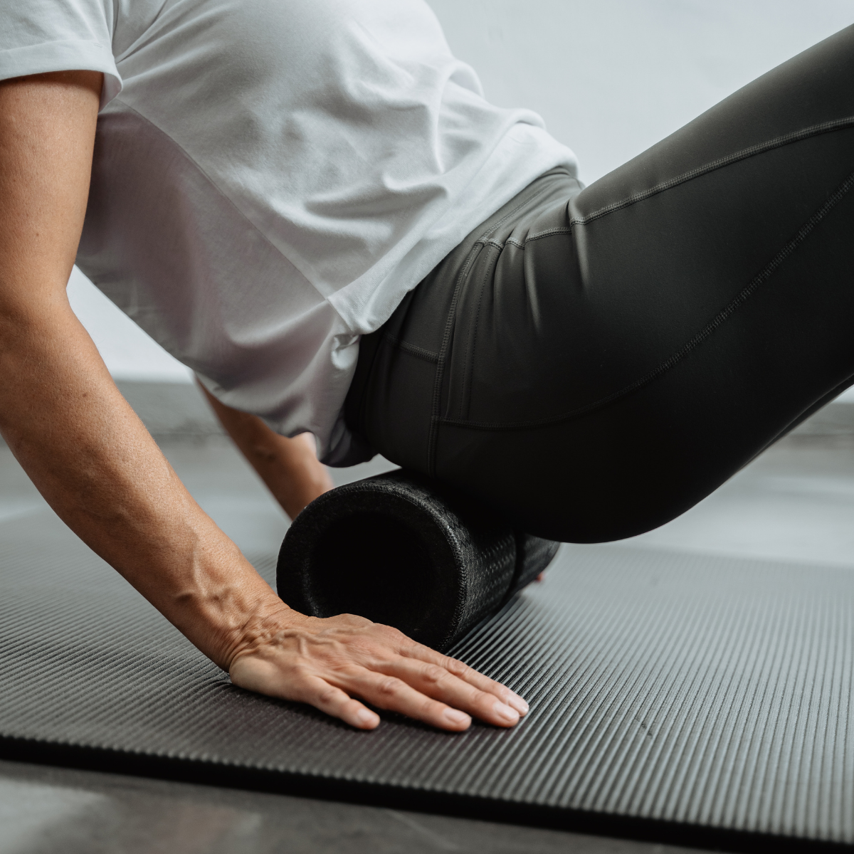 Eine Person, die ein weißes Hemd und schwarze Leggings trägt, wendet die „Liebscher & Bracht MAXI-Faszienrolle mit Wirbelsäulen-Entlastung“ auf ihrem Rücken an, während sie auf einer schwarzen Trainingsmatte liegt.