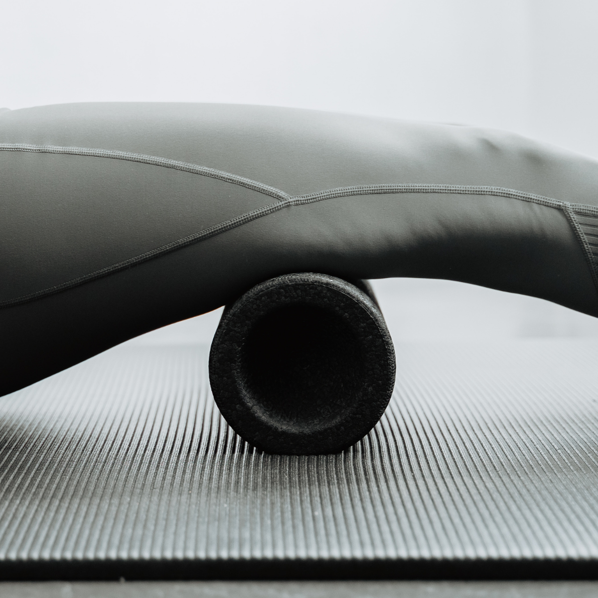 Eine Nahaufnahme des Beins einer Person in Sportbekleidung, die auf einer Yogamatte die „Liebscher & Bracht MAXI-Faszienrolle mit Wirbelsäulen-Entlastung“ verwendet, was auf eine Muskelmassage oder Physiotherapie für den Rücken hindeutet.