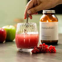 Eine Hand rührt ein rosarotes Getränk in einem strukturierten Glas um. Neben dem Glas liegen rote Johannisbeeren, im Hintergrund stehen Äpfel und ein braunes Glas mit der Aufschrift Basenpulver+.