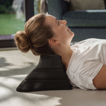 Eine Frau mit blonden Haaren, die zu einem Dutt zusammengebunden sind, liegt auf dem Boden und benutzt „Der Nackenretter“ von Liebscher & Bracht – ein ergonomisches schwarzes Schaumstoffkeil für von Liebscher & Bracht inspirierte Übungen zur Linderung von Nackenschmerzen –, während im Hintergrund Sonnenlicht und ein Sofa zu sehen sind.