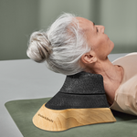 Eine ältere Frau mit grauem Haar liegt auf einer Matte und entspannt sich, während sie den Liebscher & Bracht Holz-Sockel für den Nackenretter - eine ergonomische Stütze aus schwarzem Holz - zur Ausrichtung des Nackens oder zur Therapie verwendet.