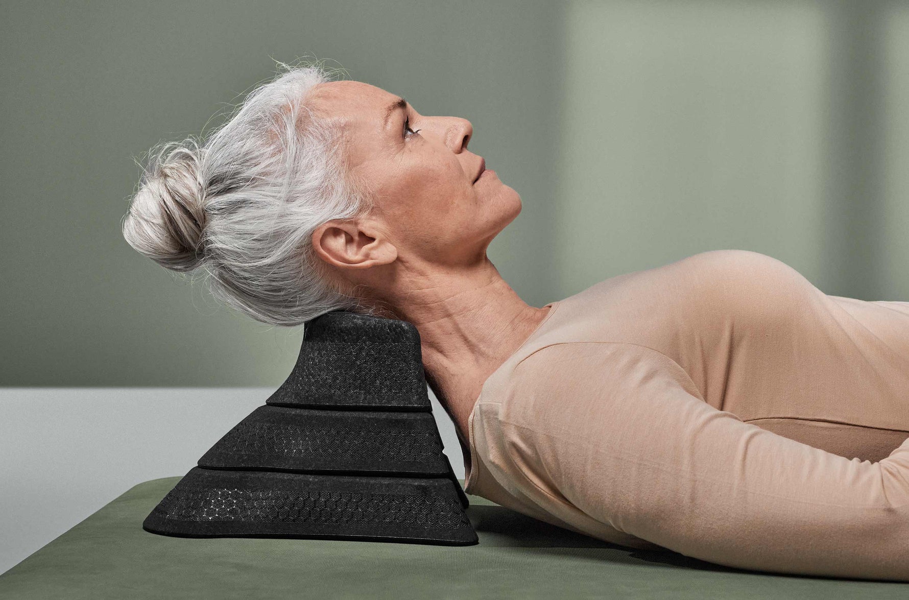 Eine ältere Frau mit grauem Haar in einem Dutt liegt auf dem Rücken und benutzt ein schwarzes Nackenstützkissen. Sie trägt ein beigefarbenes langärmeliges Hemd und ruht auf einer grünen Fläche in einer minimalistischen, modernen Umgebung.