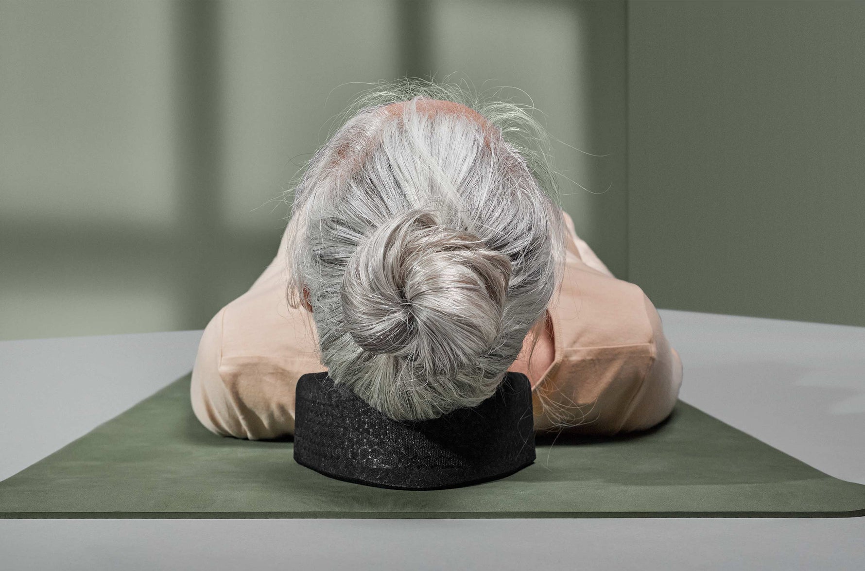 Eine ältere Person mit grauem Haar in einem Dutt liegt auf dem Rücken auf einer Matte und stützt ihren Kopf auf eine schwarze Nackenstütze, von oben gesehen. Die Umgebung ist in gedämpften Grün- und Beigetönen gehalten.