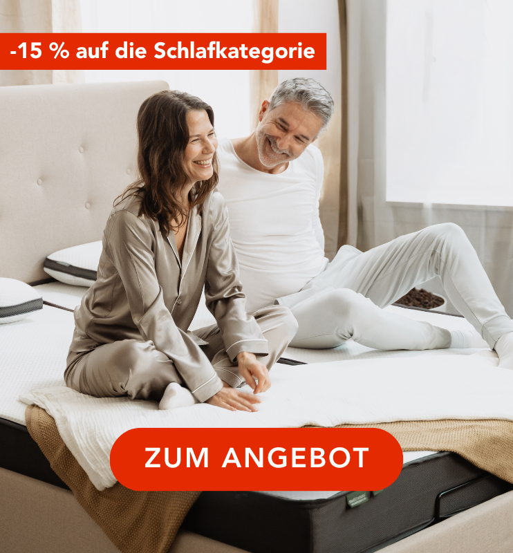 Eine lächelnde Frau und ein Mann im Schlafanzug sitzen auf einem Bett in einem hellen Schlafzimmer. Der deutsche Text lautet "-15 % auf die Schlafkategorie" und "ZUM ANGEBOT" in fetten roten Bannern.