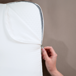 Eine Hand öffnet den Reißverschluss des weißen Liebscher & Bracht 2er-Pack: Baumwoll-Kissenbezug und gibt den Blick auf die innere Schicht vor einem neutralen, schlichten Hintergrund frei.