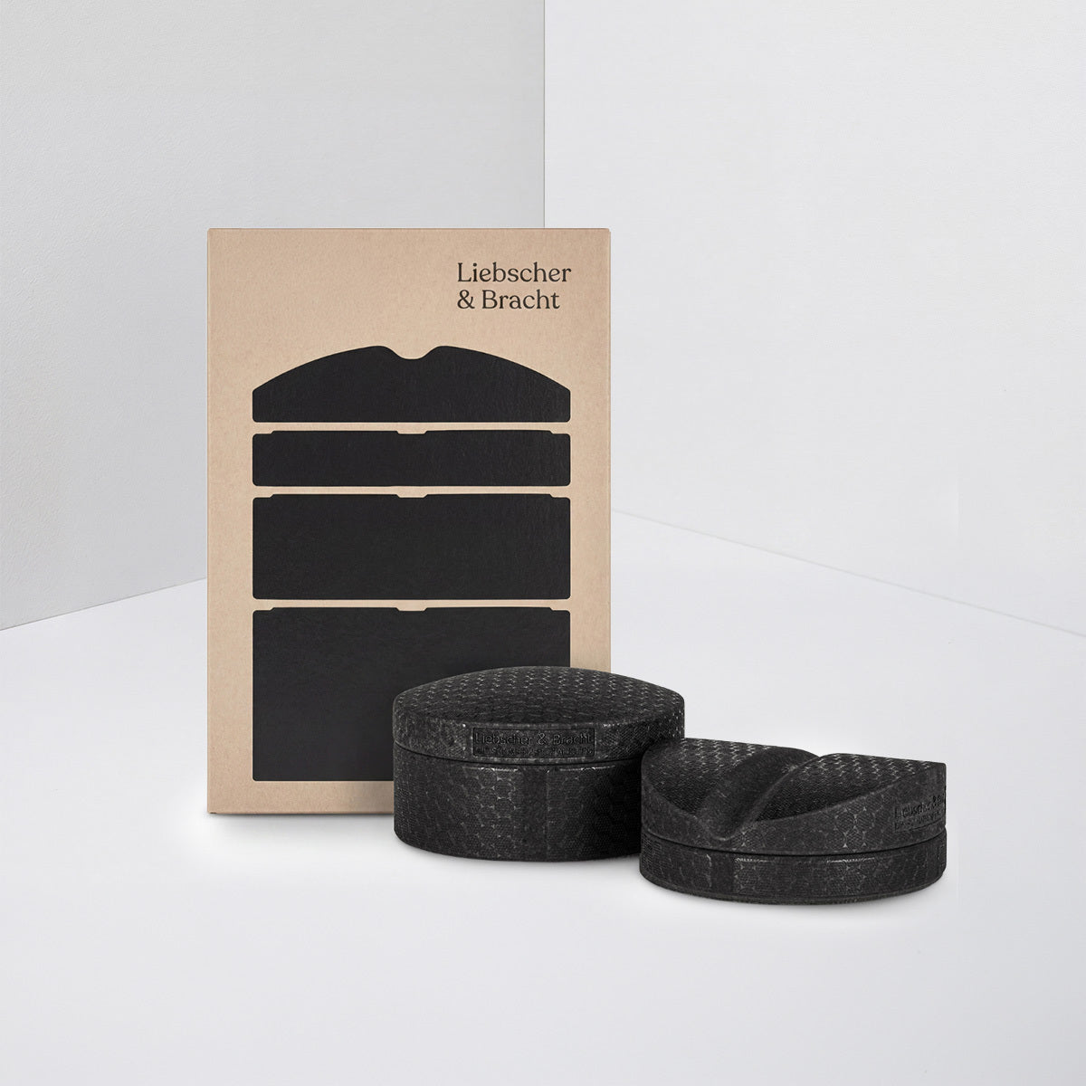 Zwei schwarze, strukturierte Schaumstoffblöcke und ein dazu passendes schwarzes Schaumstoffkissen sind vor einer braunen Verpackung mit der Aufschrift „Liebscher & Bracht“ angeordnet, die auf einer weißen Fläche mit minimalistischem Hintergrund steht. Das Set „Der Rückenretter“ von Liebscher & Bracht eignet sich ideal für Übungen gegen Rückenbeschwerden und wurde entwickelt, um Ihnen zu helfen, Ihre Haltung zu verbessern und Rückenschmerzen zu lindern.