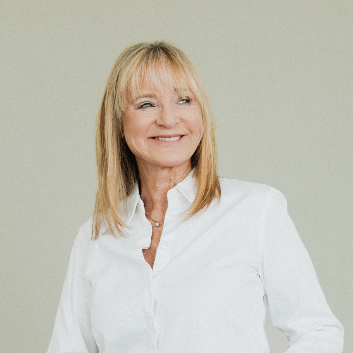 Eine lächelnde ältere Frau mit glattem blondem Haar und Pony, die ein weißes Button-up-Hemd trägt, steht vor einem schlichten hellen Hintergrund und schaut leicht nach links.