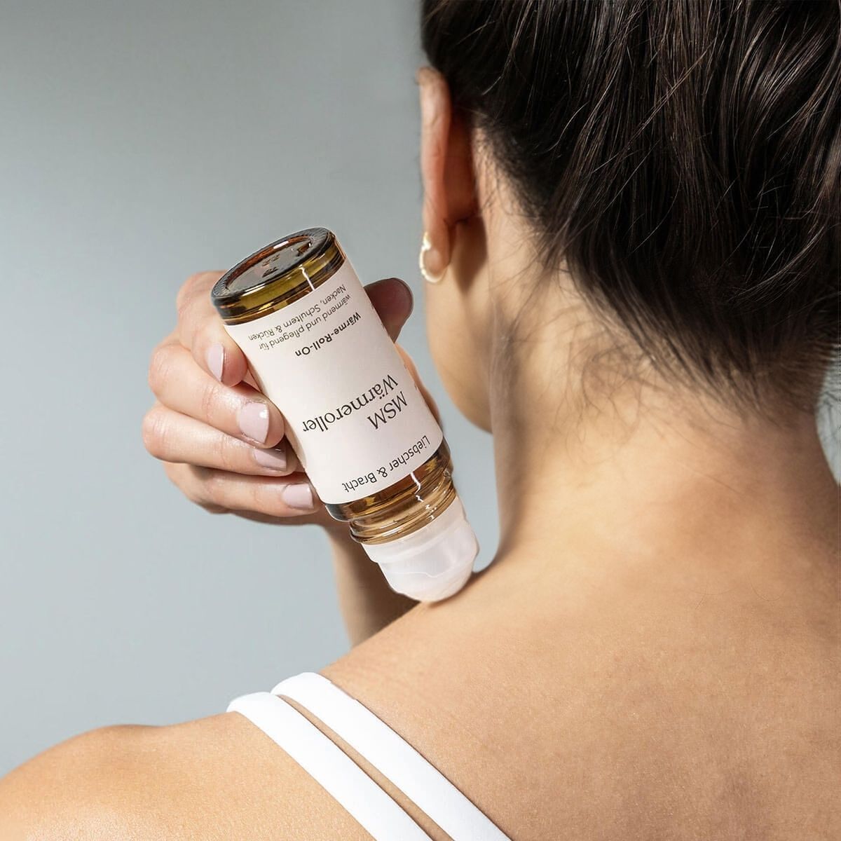 Eine Person trägt den Liebscher & Bracht MSM Wärmeroller mit natürlichen Inhaltsstoffen mit dem Rollerapplikator auf den Nacken auf. Mit zurückgebundenen dunklen Haaren und einem weißen, dünn geschnürten Oberteil genießt sie einen Moment des Wohlbefindens.