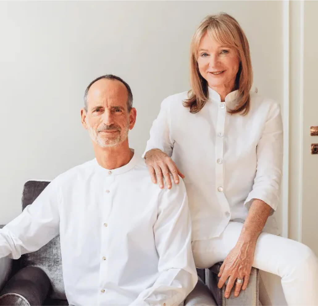 Ein Mann und eine Frau, beide hellhäutig und mit weißen Hemden bekleidet, posieren zusammen in einem Innenraum. Der Mann sitzt auf einem Stuhl, während die Frau neben ihm steht und ihre Hand auf seiner Schulter ruht. Sie lächeln sanft.