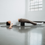 Eine Frau macht auf einer Yogamatte Übungen gegen Rückenbeschwerden für eine bessere Haltung und legt dabei „Der Rückenretter“ von Liebscher & Bracht unter ihren unteren Rücken – in einem hellen, minimalistisch eingerichteten Raum.