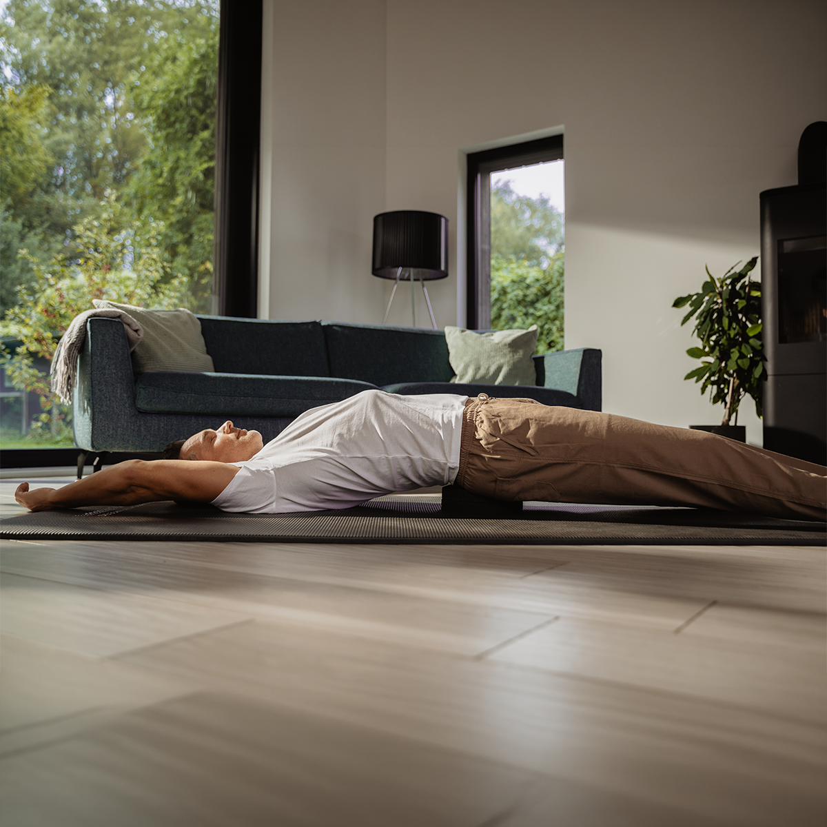 Eine Person nutzt den „Rückenretter“ von Liebscher & Bracht auf einer Yogamatte in einem Raum und streckt dabei die Arme über den Kopf und die Beine gerade aus, um Übungen gegen Rückenbeschwerden durchzuführen. Ein Sofa, ein Fenster und eine Pflanze in sanftem Tageslicht bilden einen ruhigen Hintergrund.