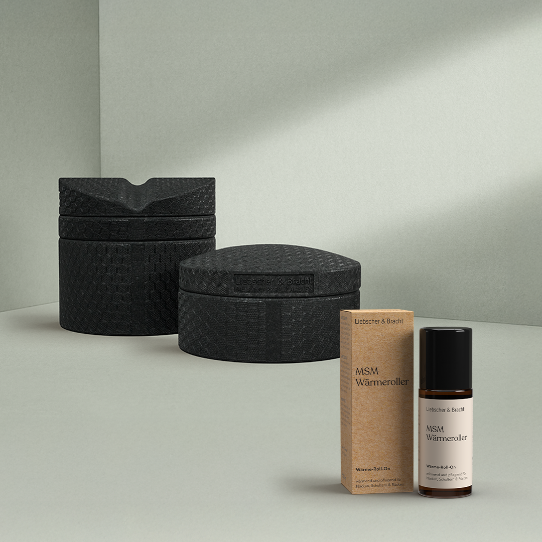 Im Vordergrund ist das Liebscher & Bracht Rückenretter + MSM-Wärmeroller-Set zu sehen - eine braune Schachtel und eine beschriftete Glasflasche mit schwarzem Verschluss -, während im Hintergrund zwei strukturierte schwarze Behälter auf einer minimalistischen Fläche stehen.