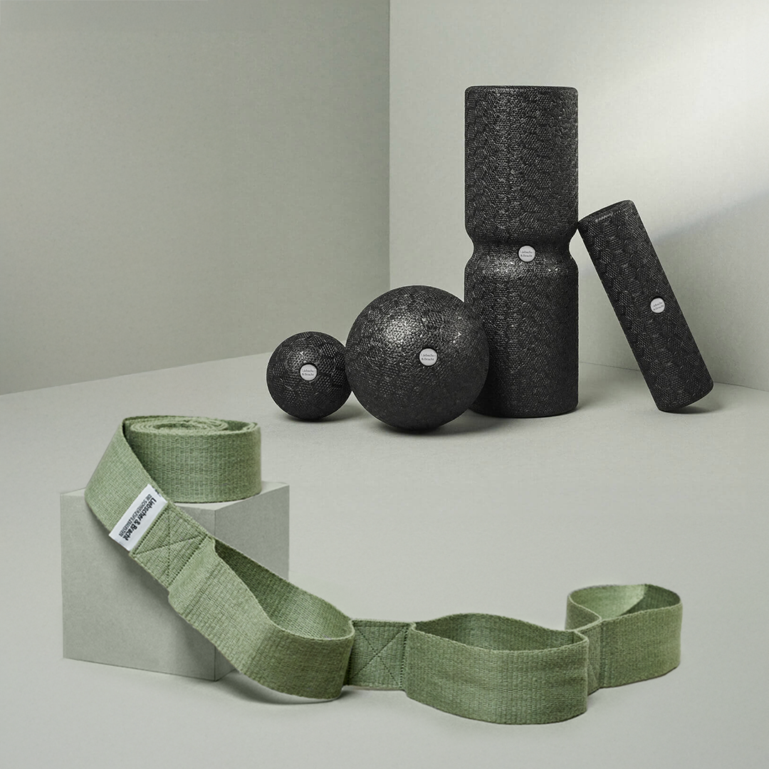 Das Liebscher & Bracht Dein Schulter-Paket zeigt schwarz strukturierte Faszienrollen und Faszienkugeln auf einer hellgrauen Oberfläche, mit einem grünen Stoff-Yogaband, das auf einem kleinen Block im Vordergrund gerollt ist.