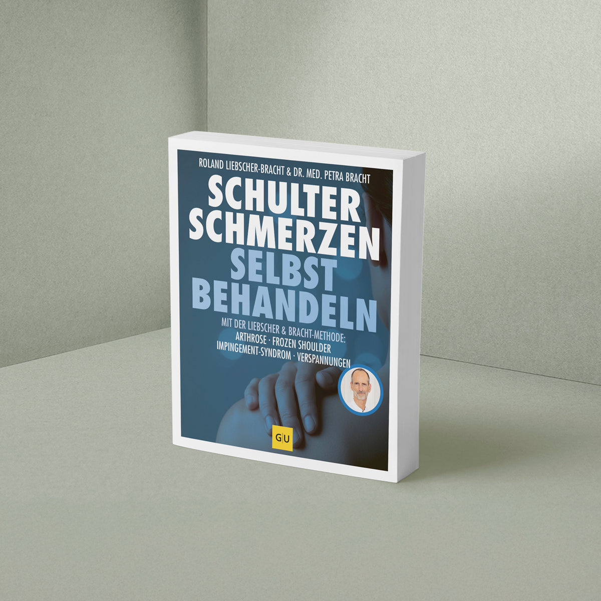 Ein Buch mit dem Titel „Schulterschmerzen selbst behandeln“ von Liebscher & Bracht steht aufrecht vor einer hellgrünen Wand und einem hellgrünen Hintergrund. Der blaue Einband ist mit weißem und blauem Text bedruckt und zeigt in der unteren rechten Ecke ein kleines rundes Foto einer Person.