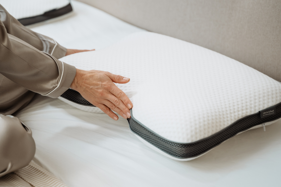 Eine Person im Schlafanzug rückt ein weißes Memory-Schaumstoffkissen mit schwarzem Netzrand zurecht, während sie auf einem ordentlich gemachten Bett sitzt.