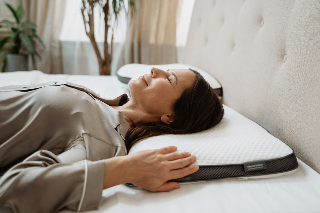 Eine Frau in einem satinierten Pyjama liegt bequem auf dem Rücken auf einem Bett, lächelt mit geschlossenen Augen und stützt ihre Hand auf ein weißes, strukturiertes Kissen. Das Sonnenlicht fällt durch die Vorhänge und im Hintergrund sind Pflanzen zu sehen.
